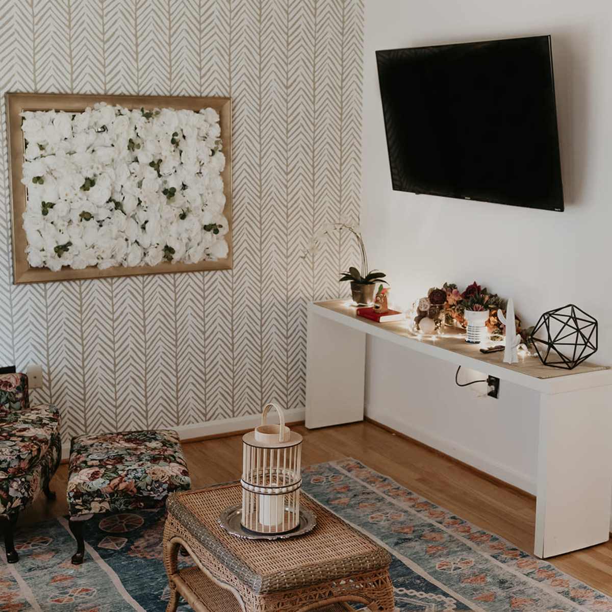 A cozy living room with floral-patterned furniture, a wall-mounted TV, and a console table decorated with plants and decorative objects.