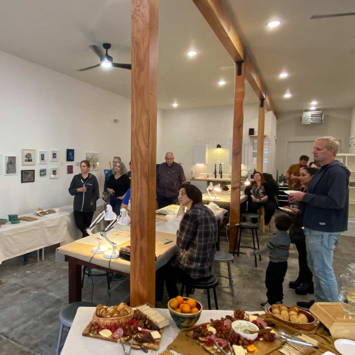 A woman stands and speaks to a small group in a modern gathering space with artwork on the walls and a table laden with food.