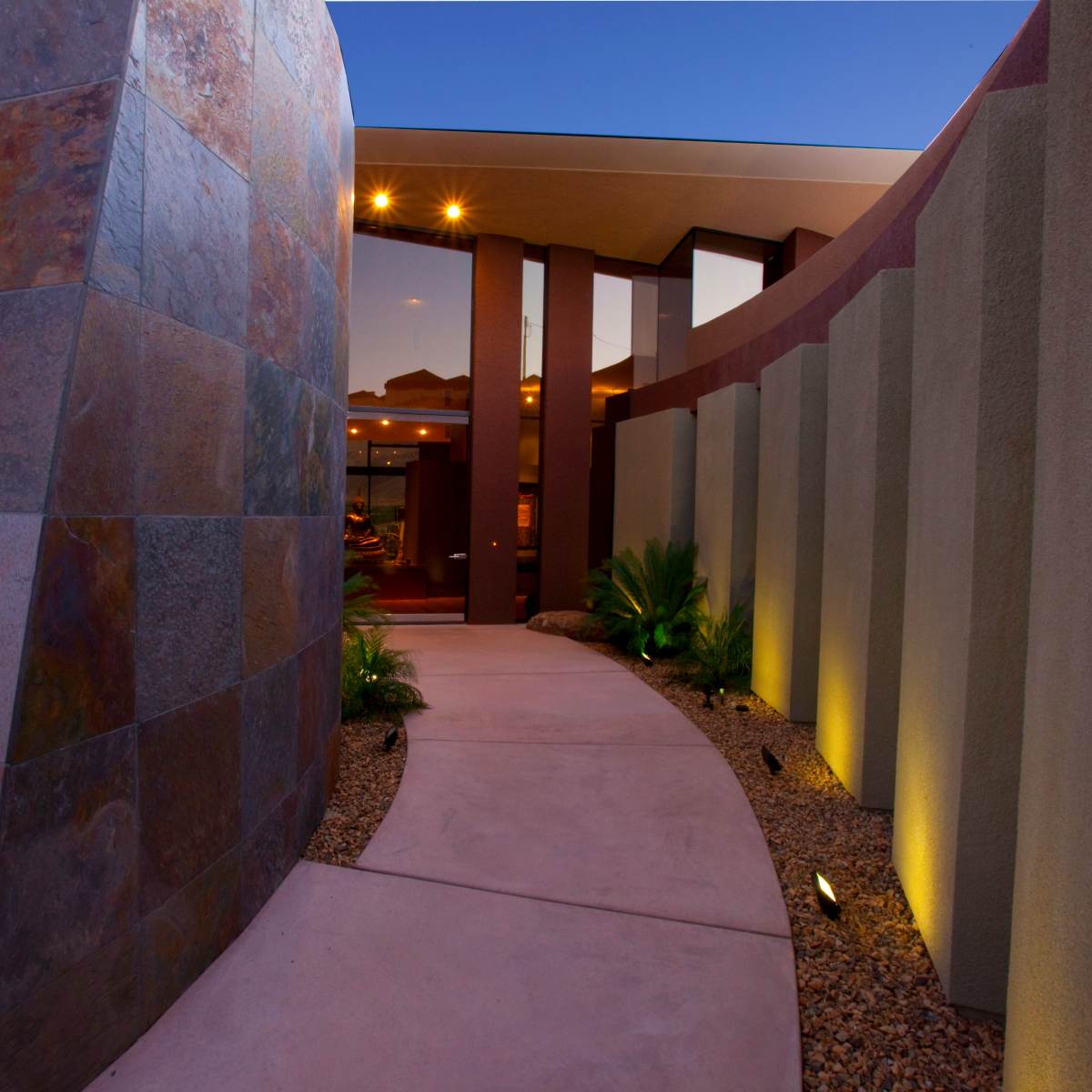 A curved pathway leading to a modern entrance surrounded by textured walls and landscaped lighting.
