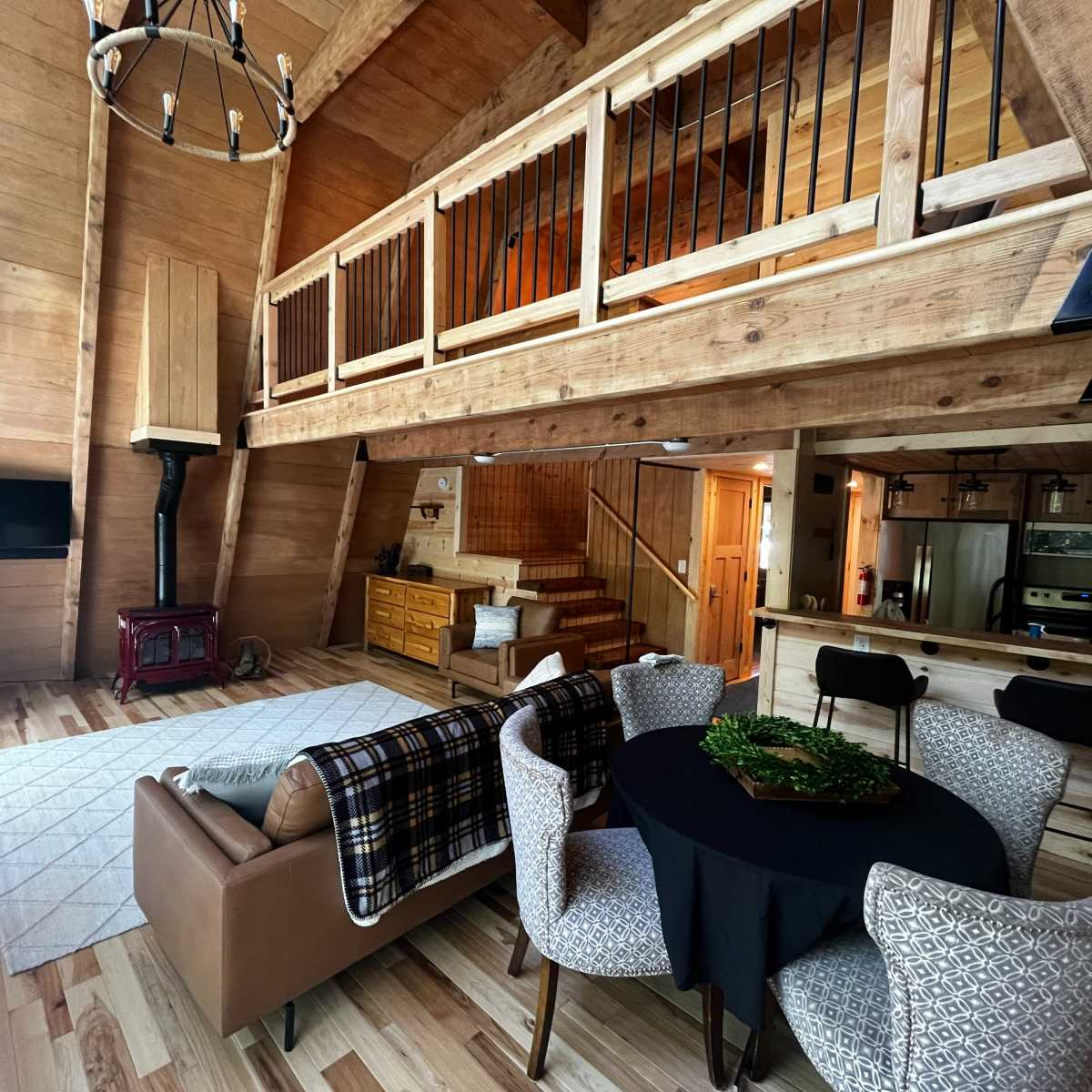 A wooden interior of a cabin featuring a loft area, a dining table, a sofa, and a staircase leading to the upper level.