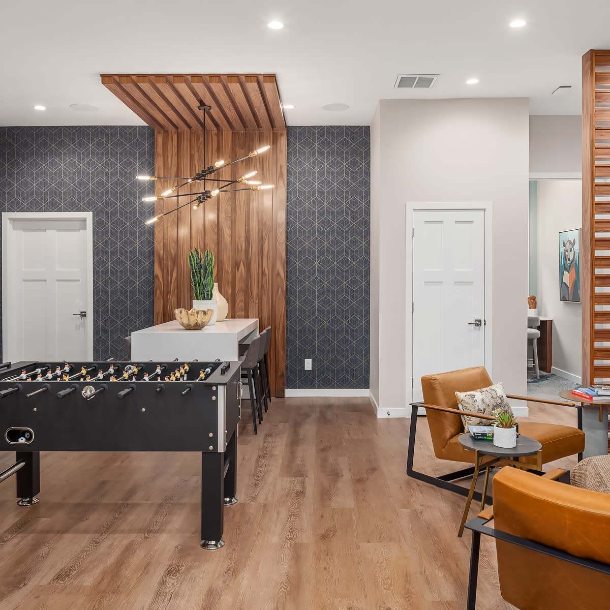 A modern lounge area featuring a foosball table, a dining setup, and cozy seating, with decorative lighting and wall patterns.