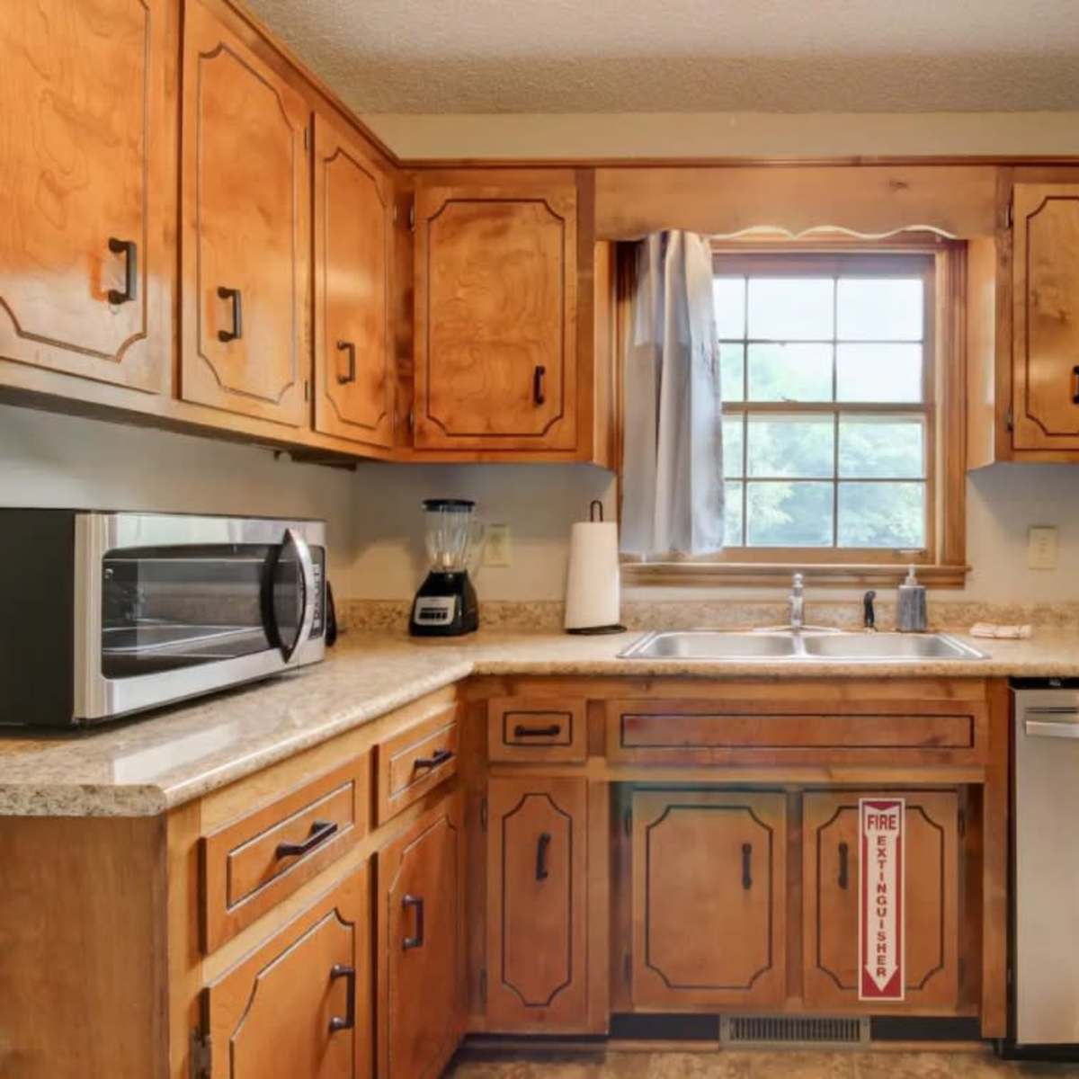 A kitchen with wooden cabinets, a stainless steel dishwasher, a sink beneath a window, and a microwave on the countertop.