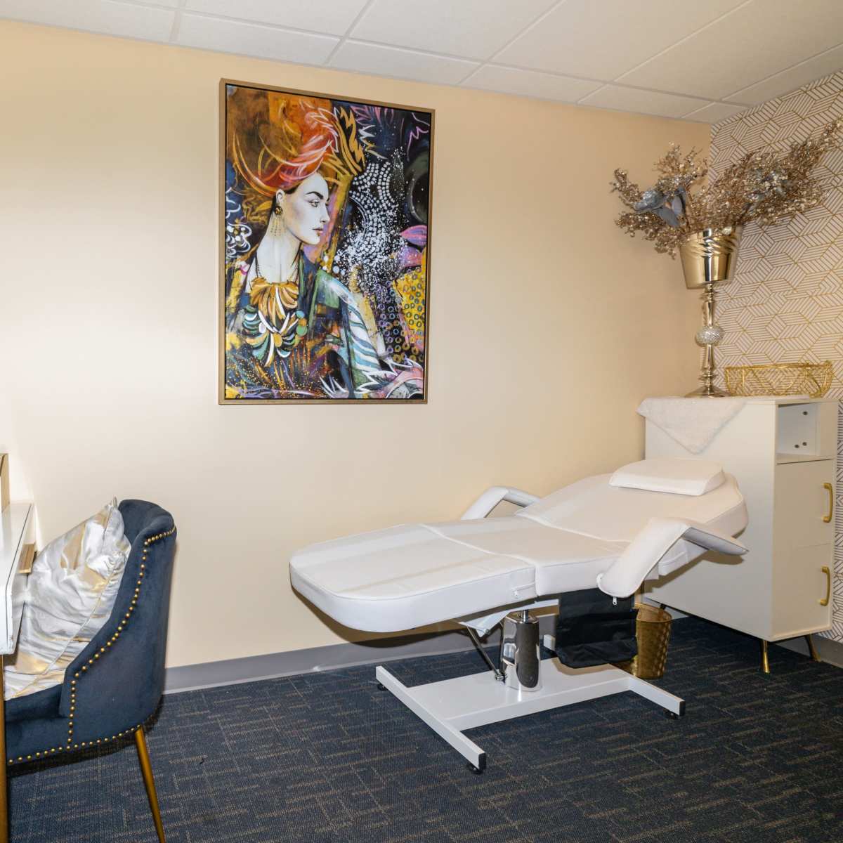 A treatment room featuring a massage table, a small desk with two blue chairs, and a colorful artwork on the wall.