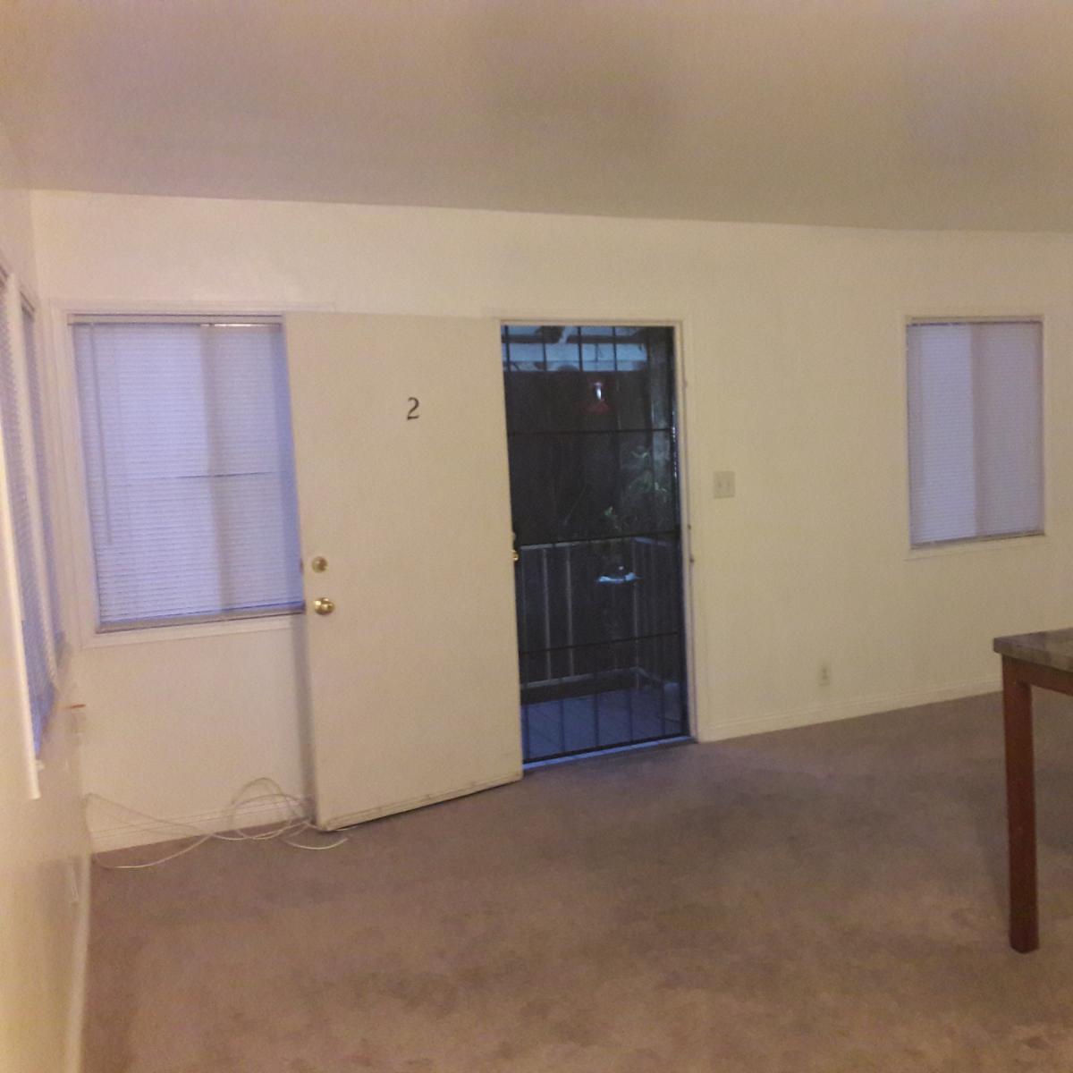 A sparsely furnished room with a door leading outside, a table in the center, and two windows on adjacent walls.