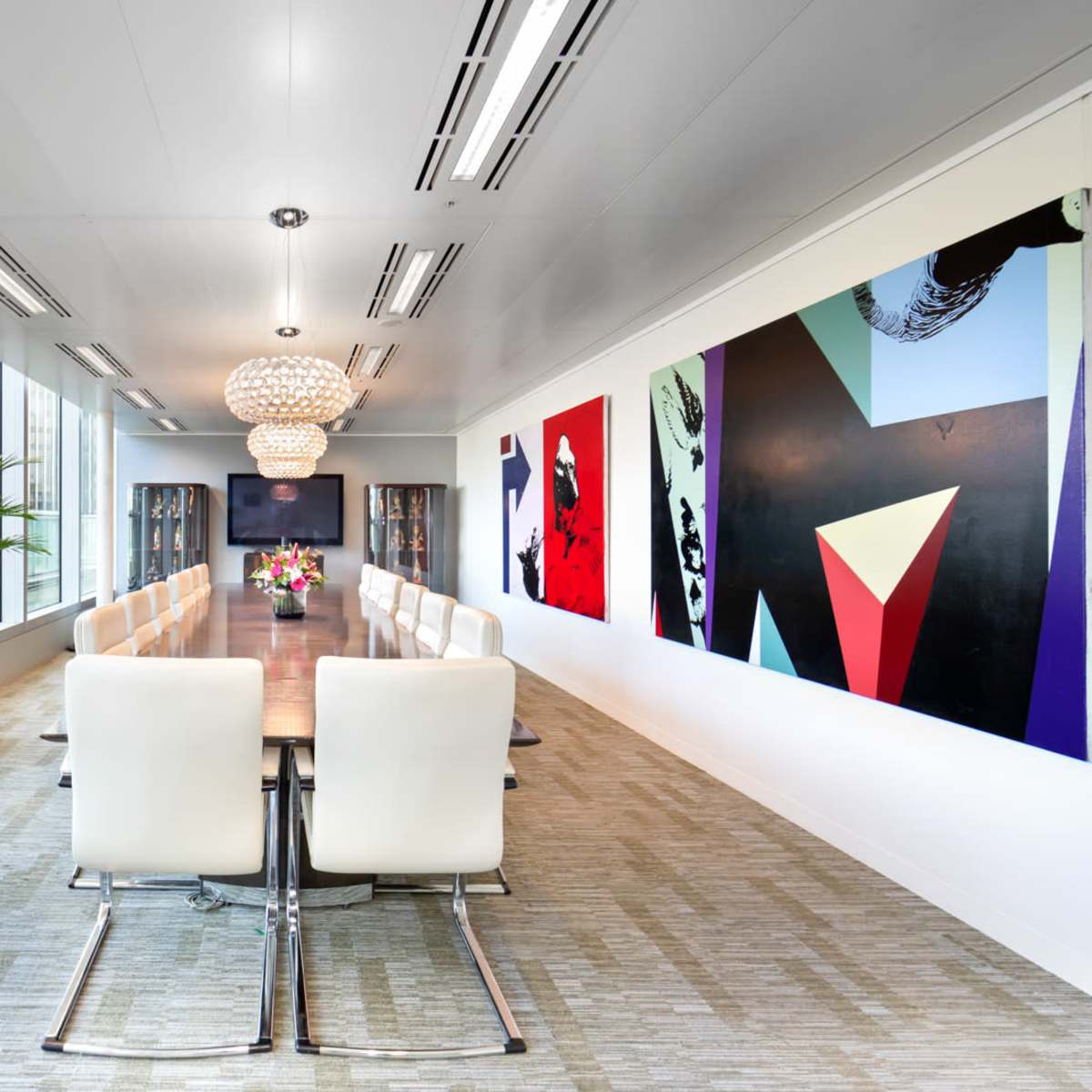 A long conference room features a large table surrounded by white chairs, with floor-to-ceiling windows on one side and colorful artwork on the walls.