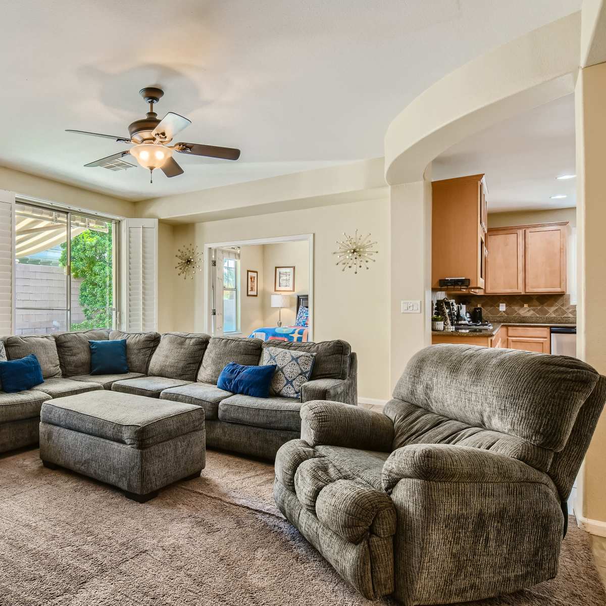 A spacious living room features a large sectional sofa, a coffee table, and a ceiling fan, with light from windows illuminating the space.