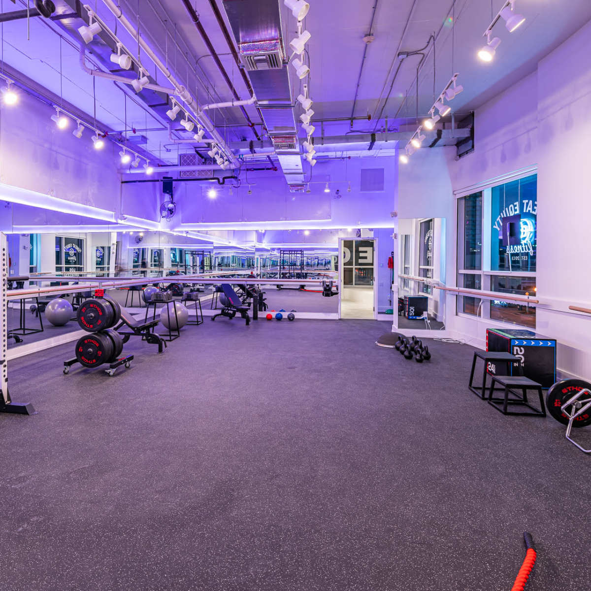 A spacious, modern gym with various weightlifting equipment, exercise mats, and mirrors under purple lighting.