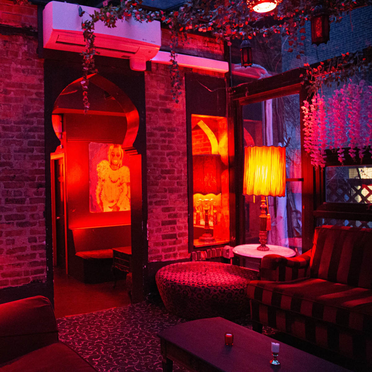 A cozy lounge features brick walls, red lighting, and stylish furniture, including patterned sofas and decorative lamps.