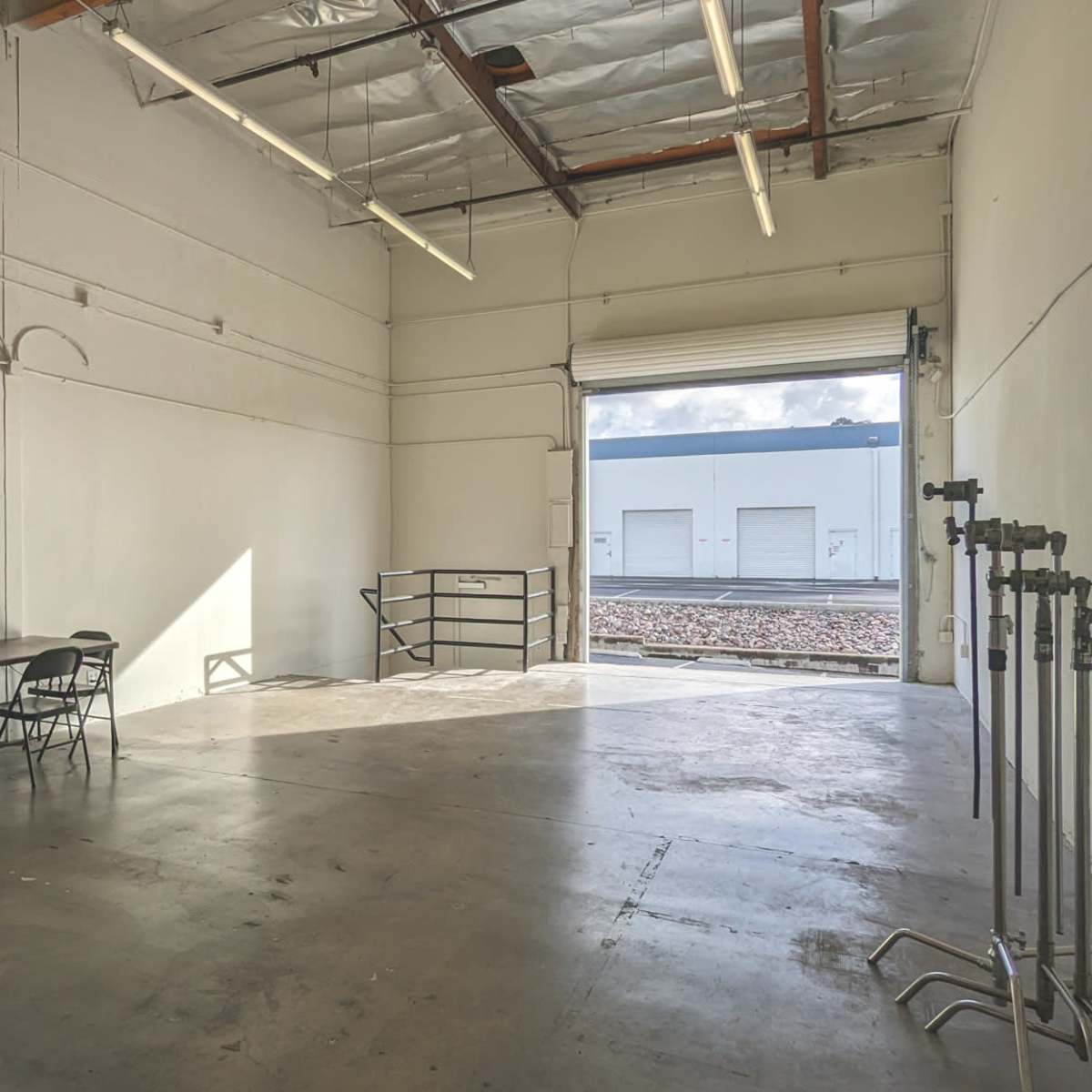 An empty industrial space with a table and chairs, a large roll-up door open to a loading dock, and a concrete floor.