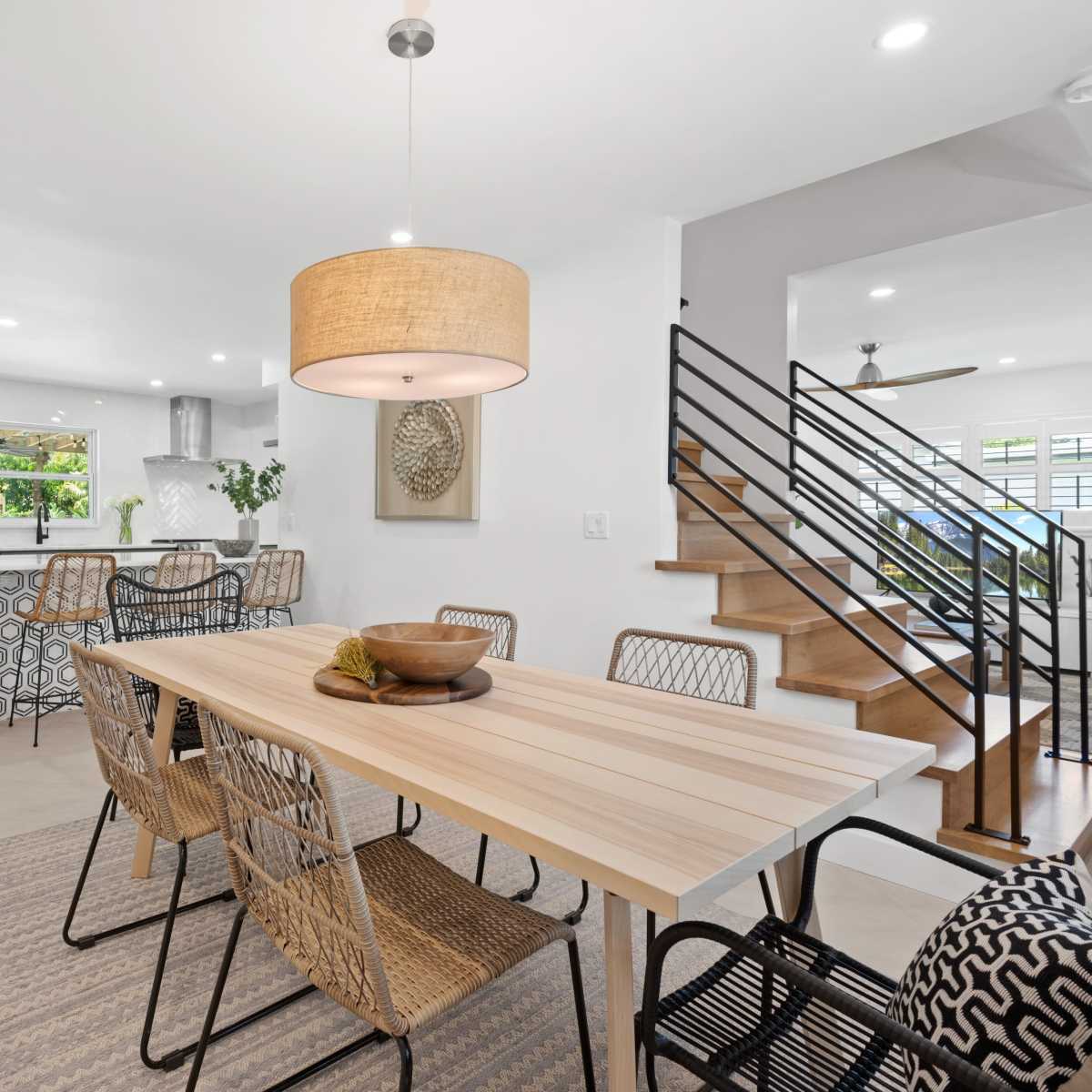 A modern dining area features a wooden table surrounded by woven chairs, with a pendant light above and a glimpse of a staircase in the background.