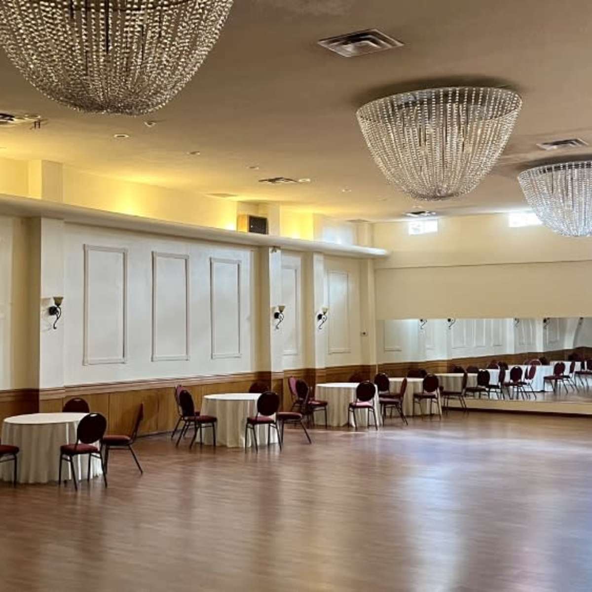 A spacious event hall with round tables and elegant chandeliers, featuring a large mirror on one wall.