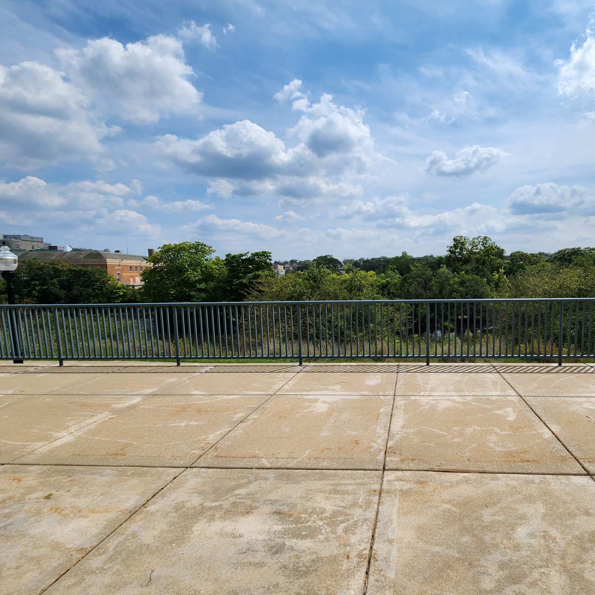 A paved area in the foreground with a railing overlooking a green landscape and blue sky with scattered clouds in the background.