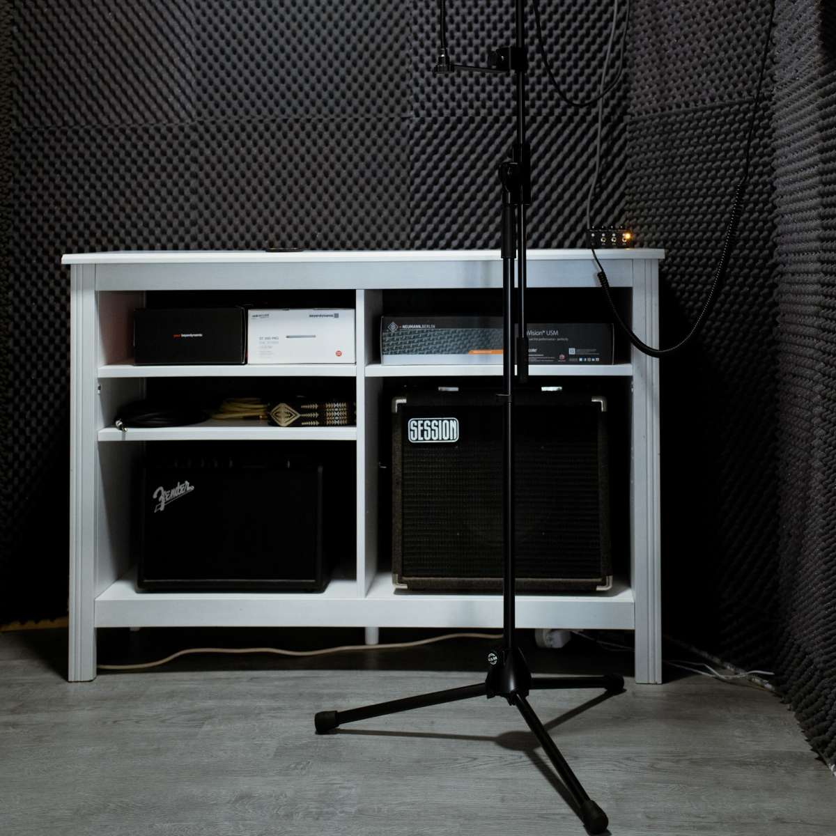 A microphone stand is positioned in front of a white shelf filled with audio equipment, all set within a soundproof room lined with black acoustic panels.