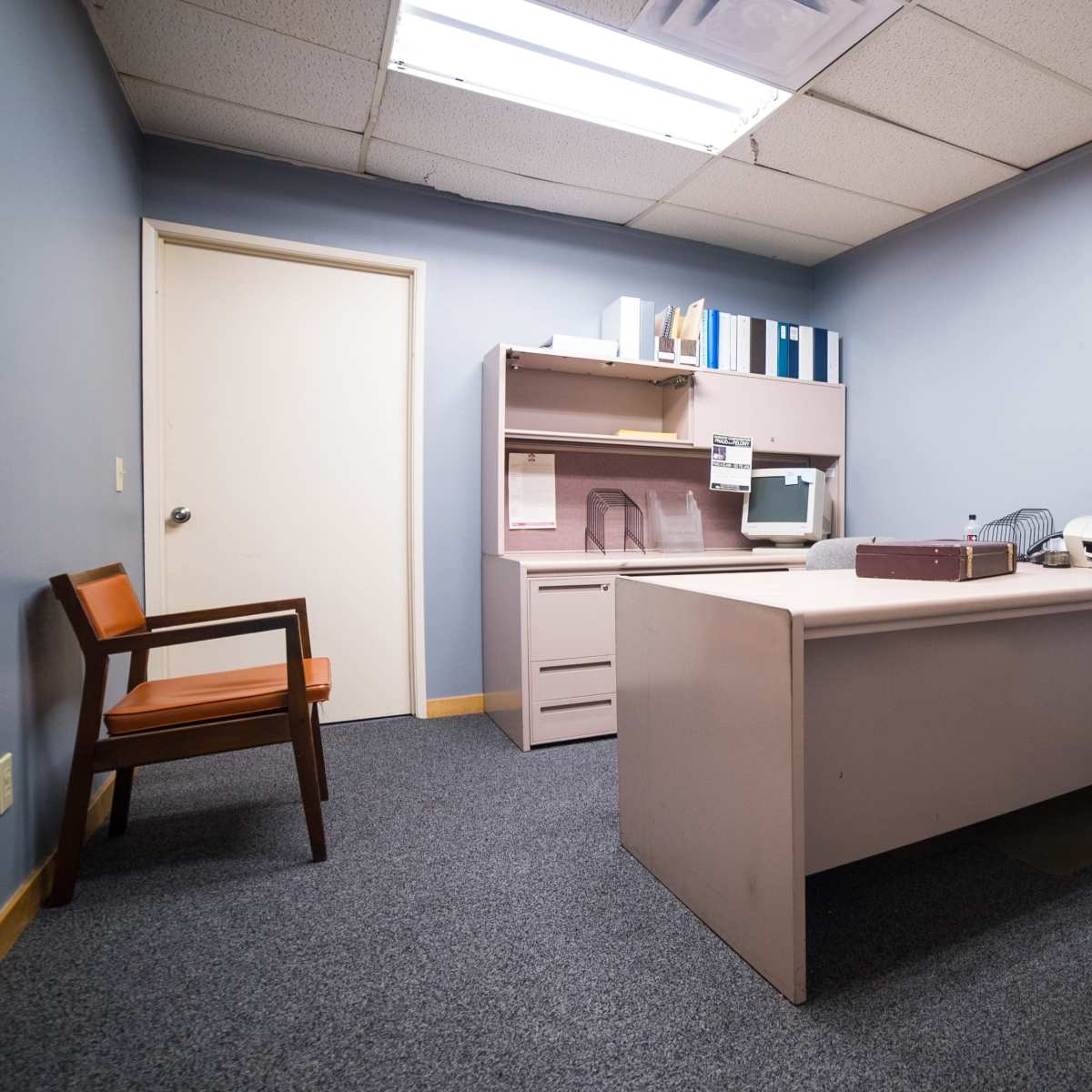 A sparsely furnished office with a desk, an old computer, and two chairs in a room with gray walls and carpeted flooring.