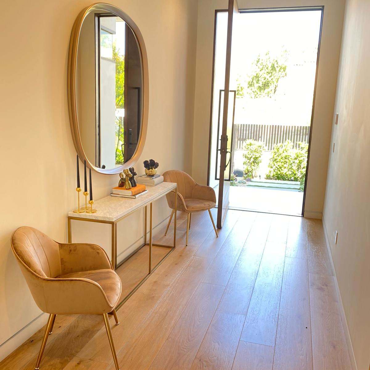A modern entryway with two stylish chairs, a console table, and a large round mirror, leading to a glass door that opens to a garden.