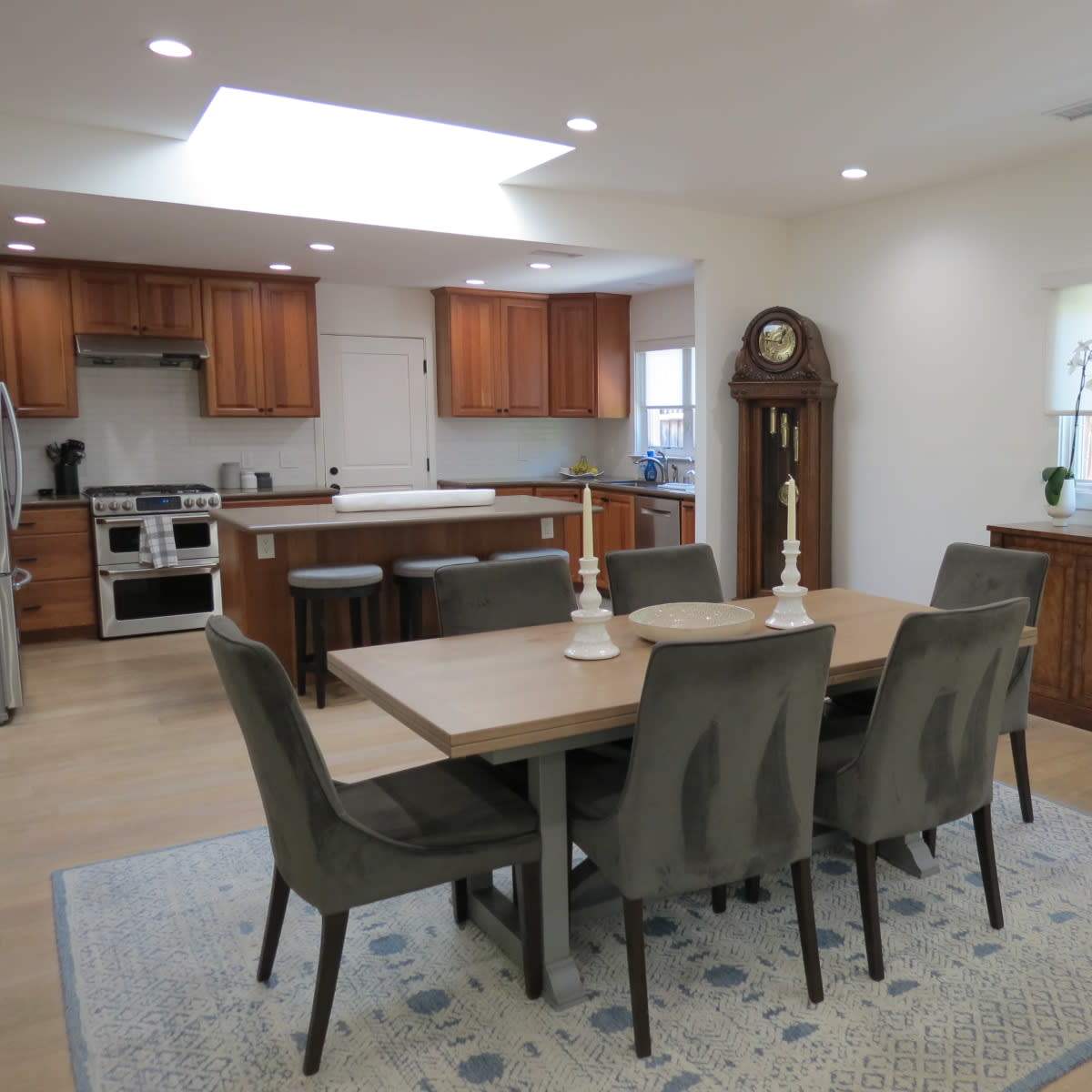 A modern kitchen and dining area with a wooden dining table, gray upholstered chairs, and a large window letting in natural light.