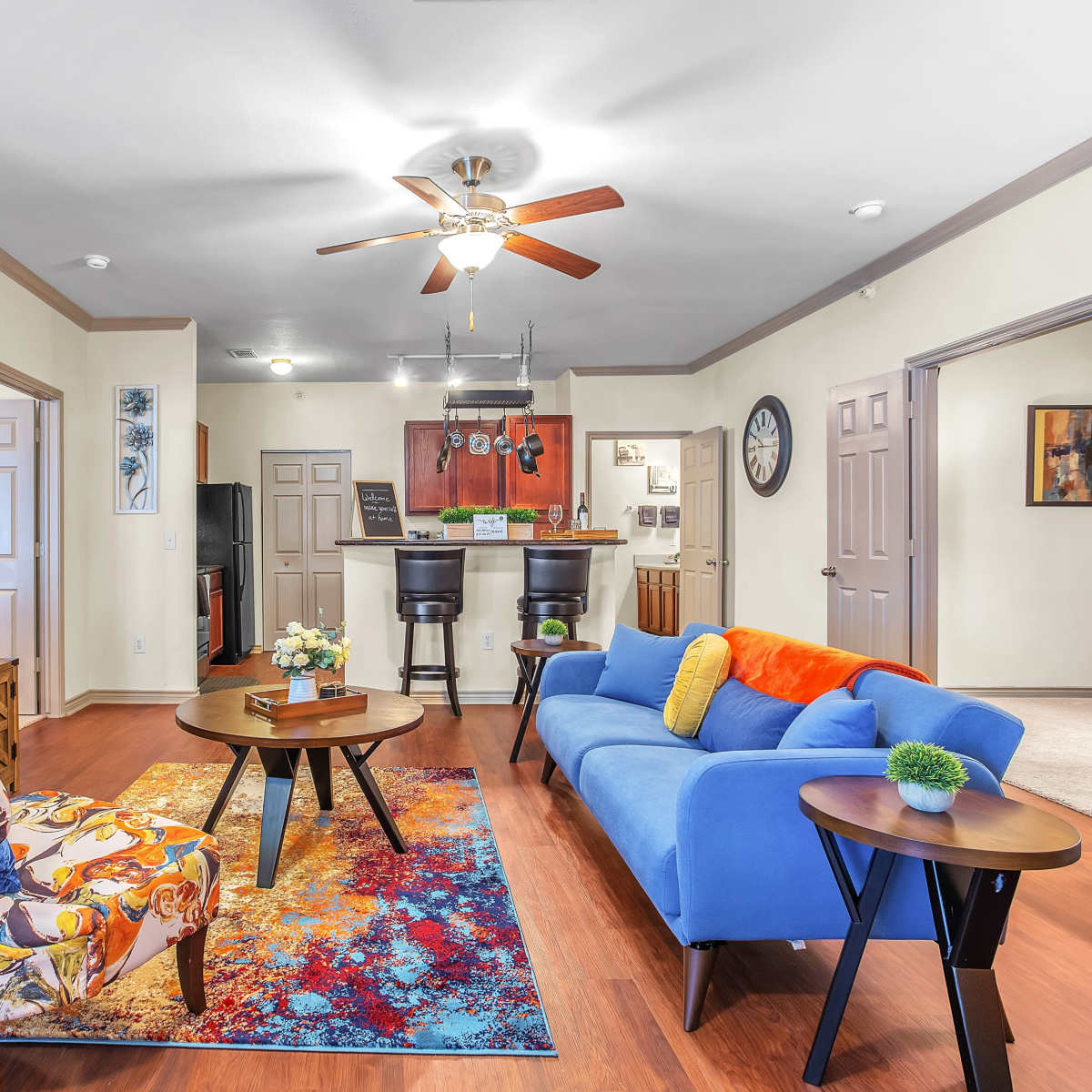 A living room with a blue sofa, an armchair with a colorful pattern, a round coffee table, and a dining area in the background featuring bar stools.