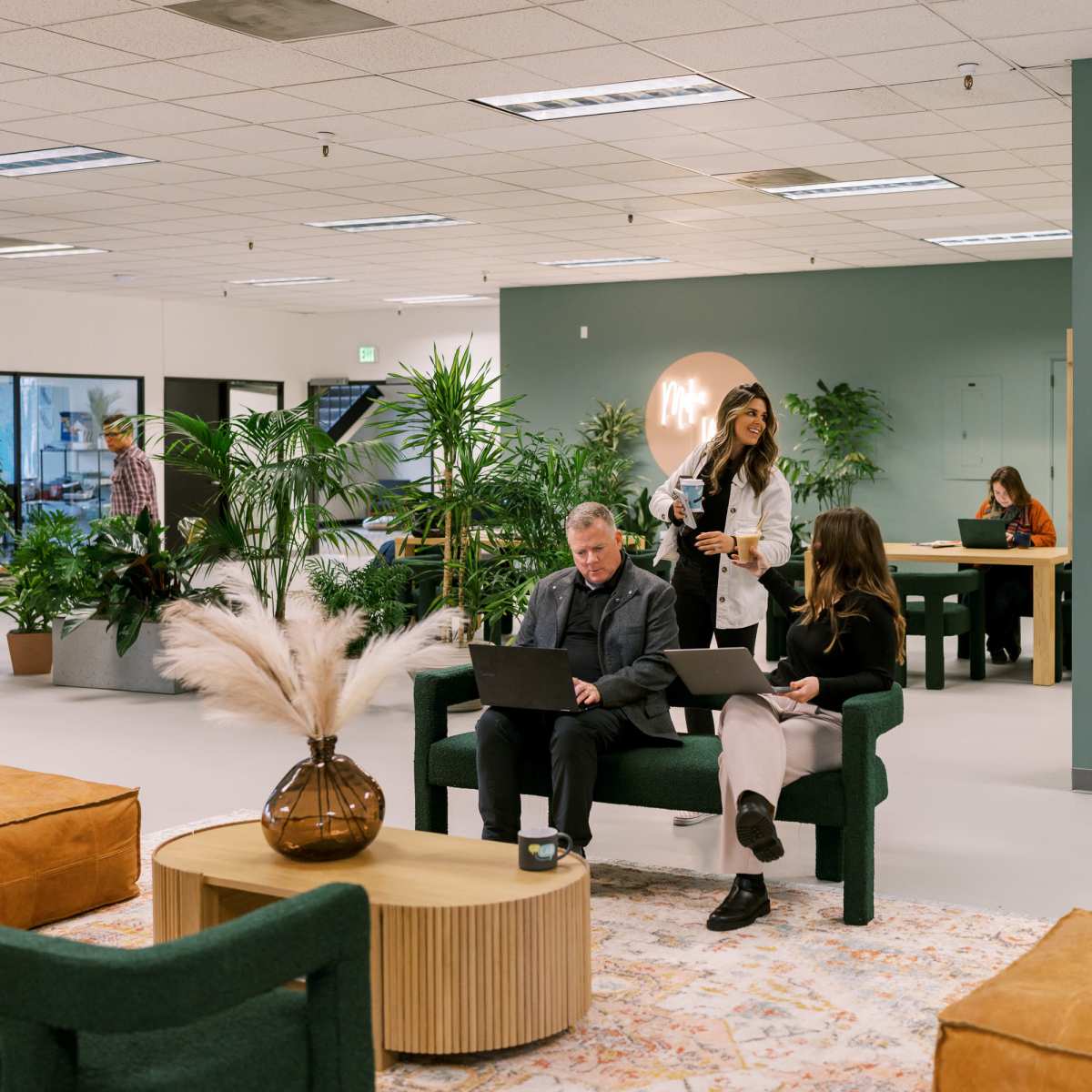 A modern office space features people working on laptops while sitting on green couches and at a communal table surrounded by plants.