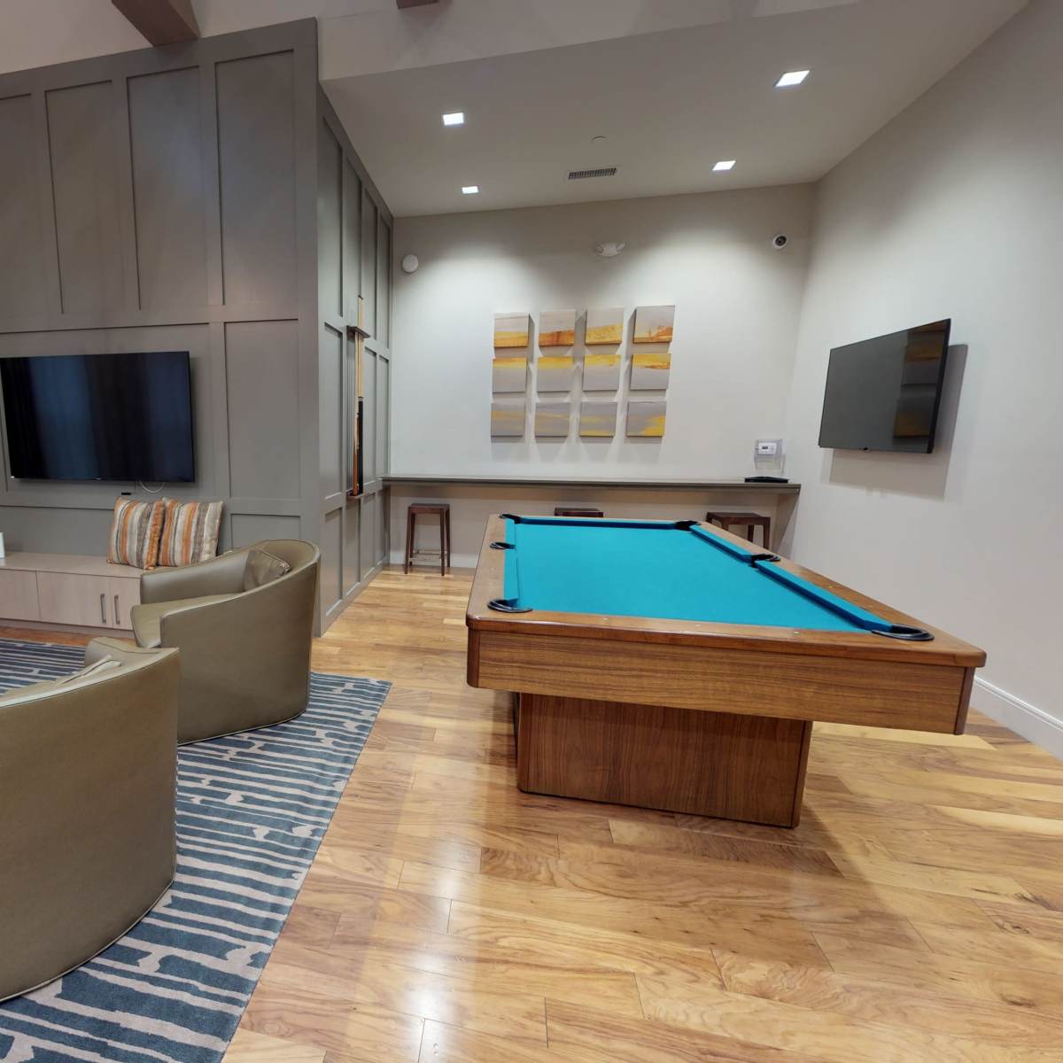 A modern living space featuring a billiards table, two armchairs, a wooden coffee table, and multiple wall-mounted televisions.