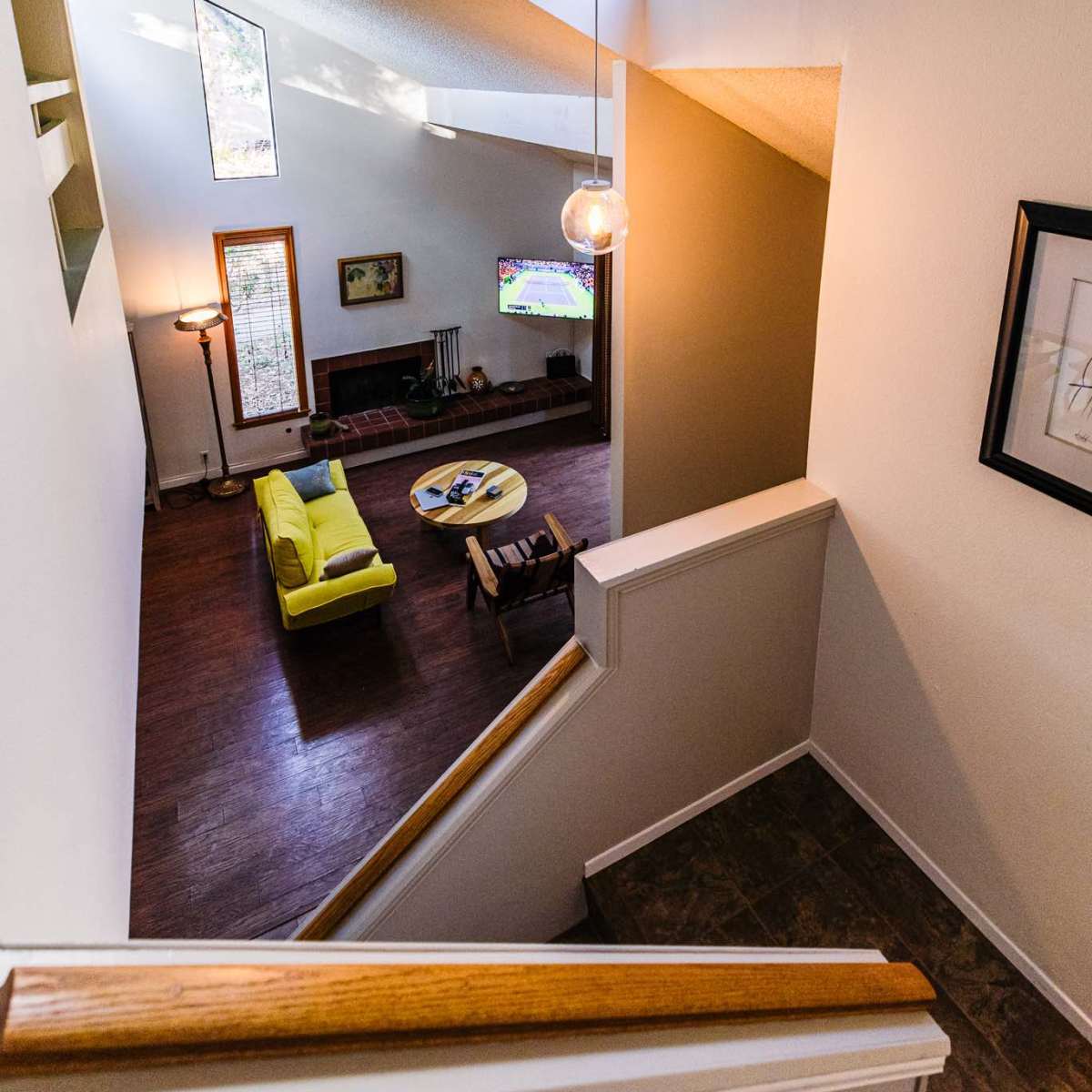 A staircase leads down to a living room with a yellow sofa and coffee table, illuminated by natural light from windows.
