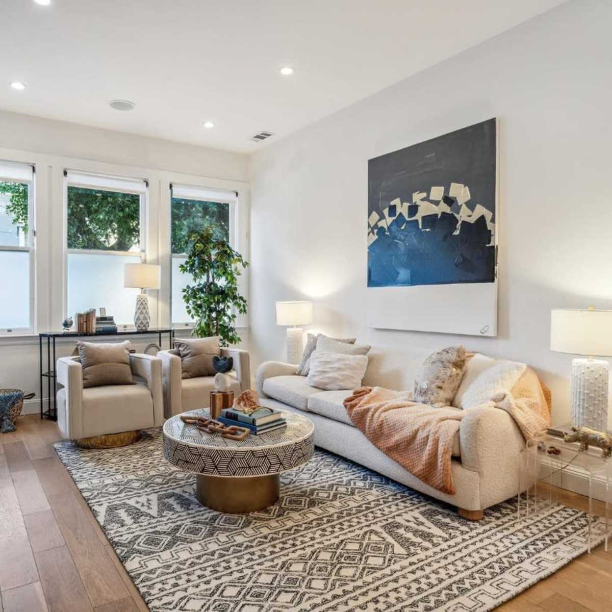 A modern living room featuring a white sofa, two armchairs, a round coffee table, and large windows allowing natural light to enter the space.