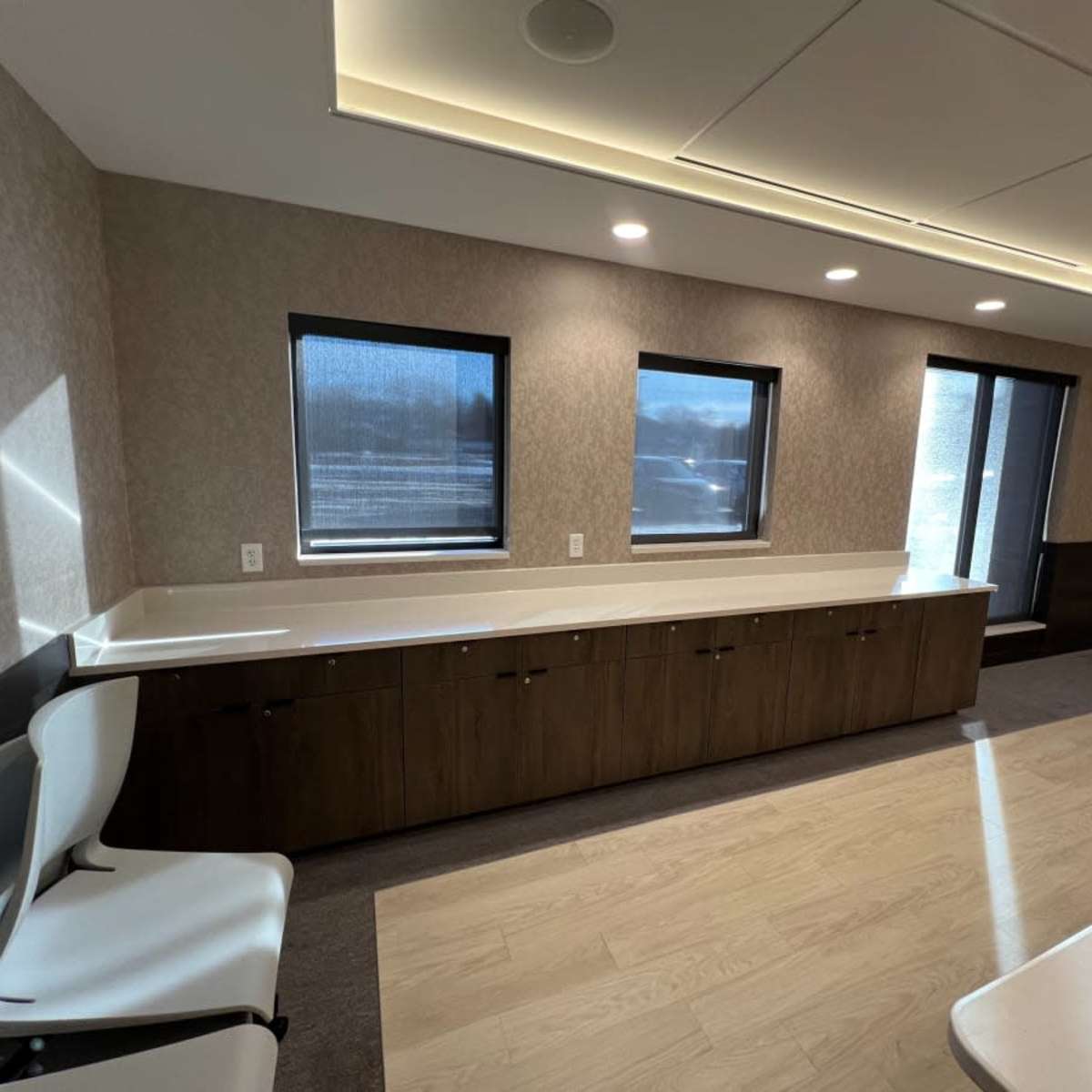 A well-lit office space featuring a long countertop with dark wood cabinetry beneath, complemented by several windows along the wall.