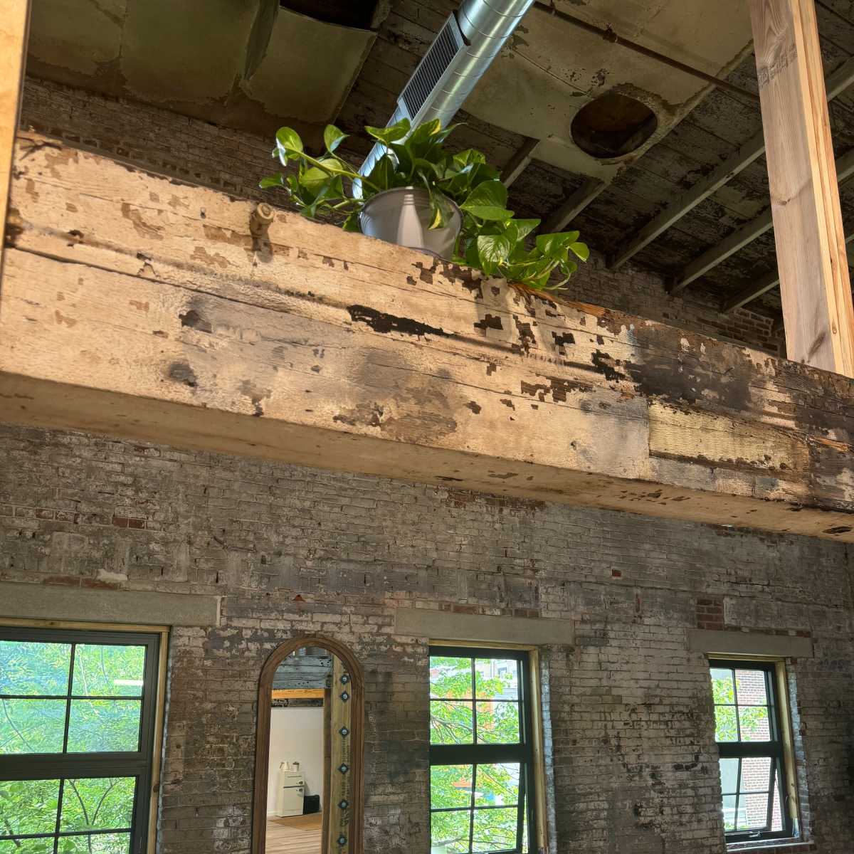A rustic interior with exposed brick walls, wooden beams, large windows, and a potted plant on a ledge.