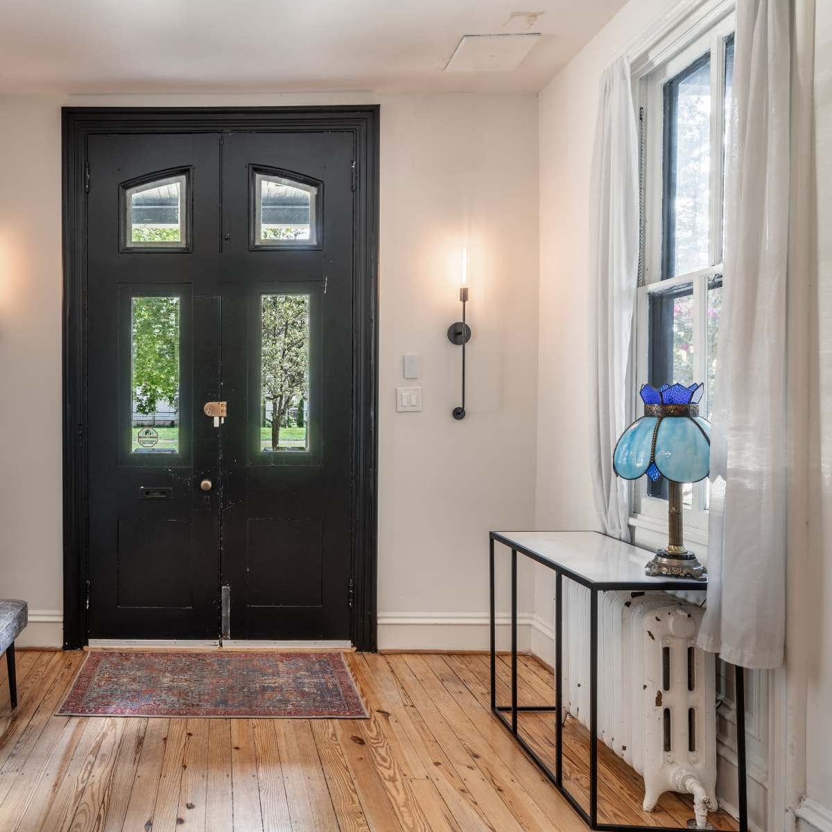 A foyer with a black double door, a bench, a lamp on a table, windows on either side, and wooden flooring.