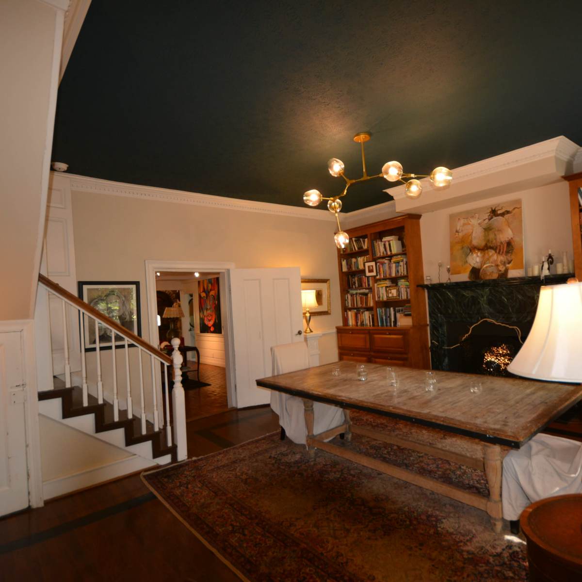 A spacious living room with a large wooden table, bookshelves filled with books, and a staircase leading to an upper level, all under a dark ceiling.