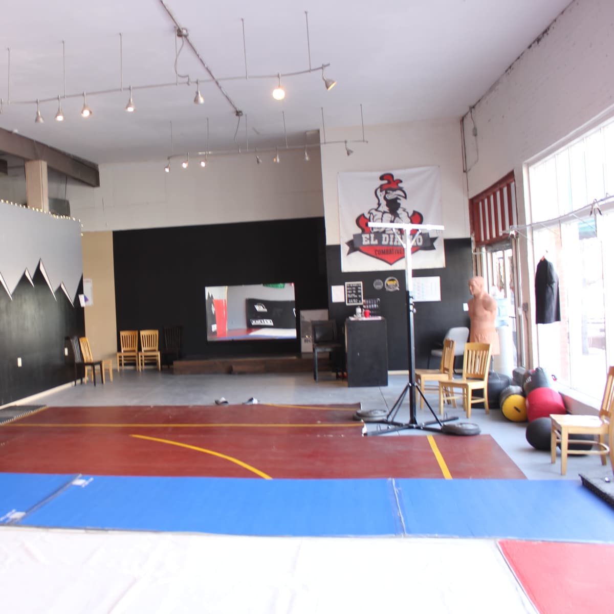 A spacious martial arts studio with a mix of wooden and padded flooring, black walls featuring a mountain design, and exercise equipment arranged near a large window.
