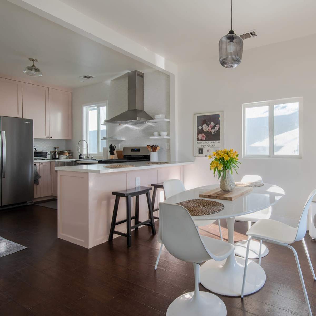 A modern kitchen and dining area features a round table with white chairs, a stainless steel refrigerator, and a large window allowing natural light to enter.