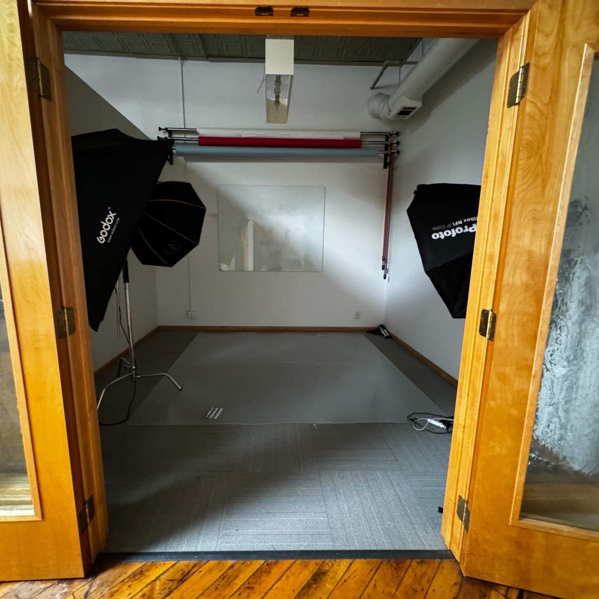 An empty studio space with wooden doors open, featuring a gray floor and two photography light stands positioned in the corners.