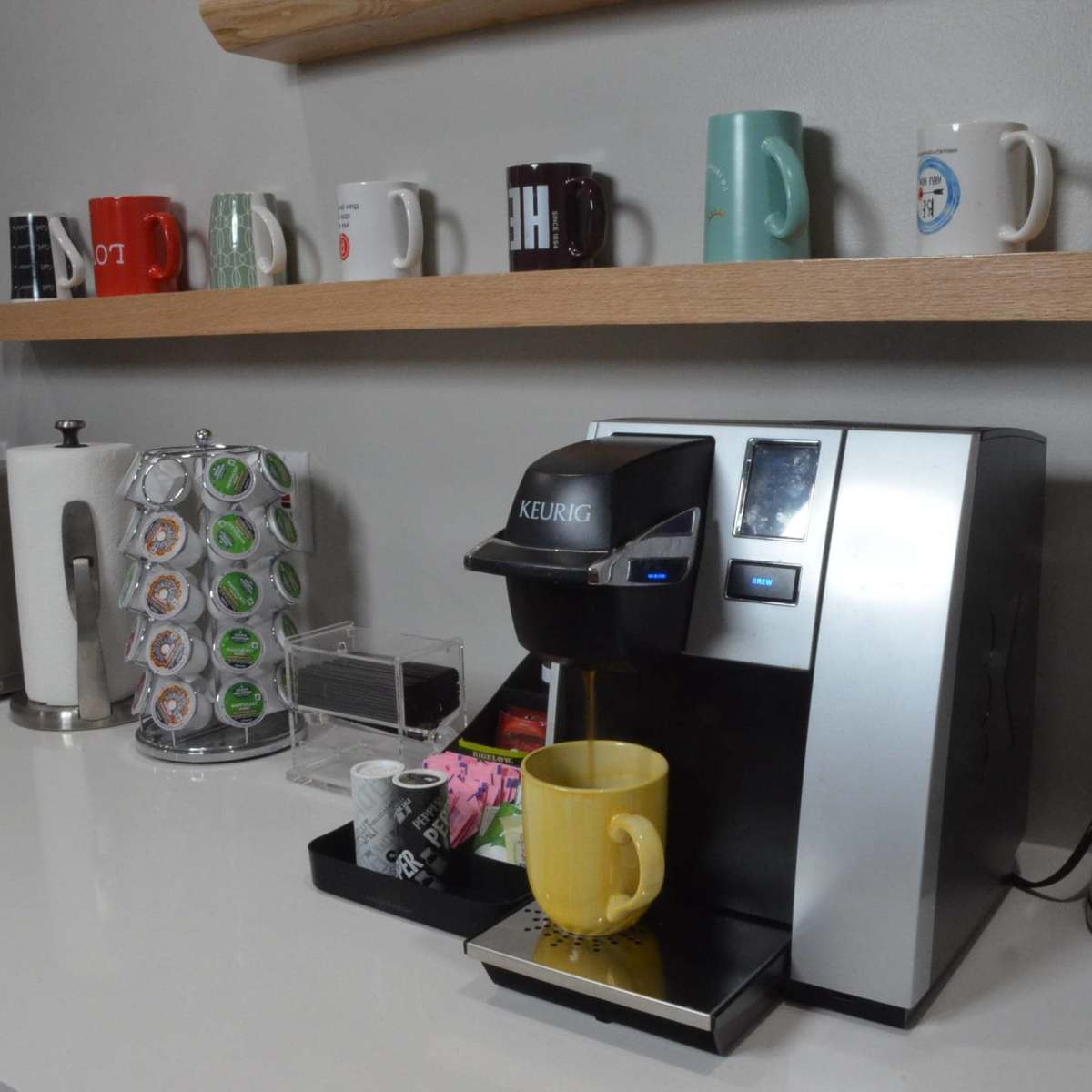 A Keurig coffee maker is positioned on a countertop with a yellow mug under the spout, surrounded by various coffee pods and a row of colorful mugs on a shelf above.