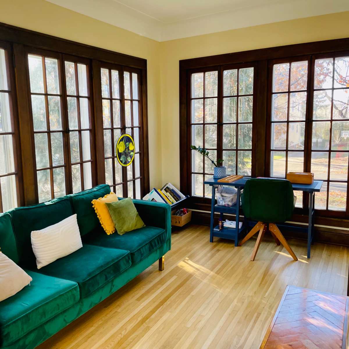 A bright room features a green sofa with colored pillows, a desk, and large windows that allow natural light to fill the space.