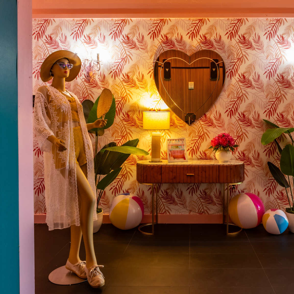 A mannequin dressed in a white outfit stands beside a wooden table adorned with a lamp and flowers, set against a backdrop of leaf-patterned wallpaper and beach balls.