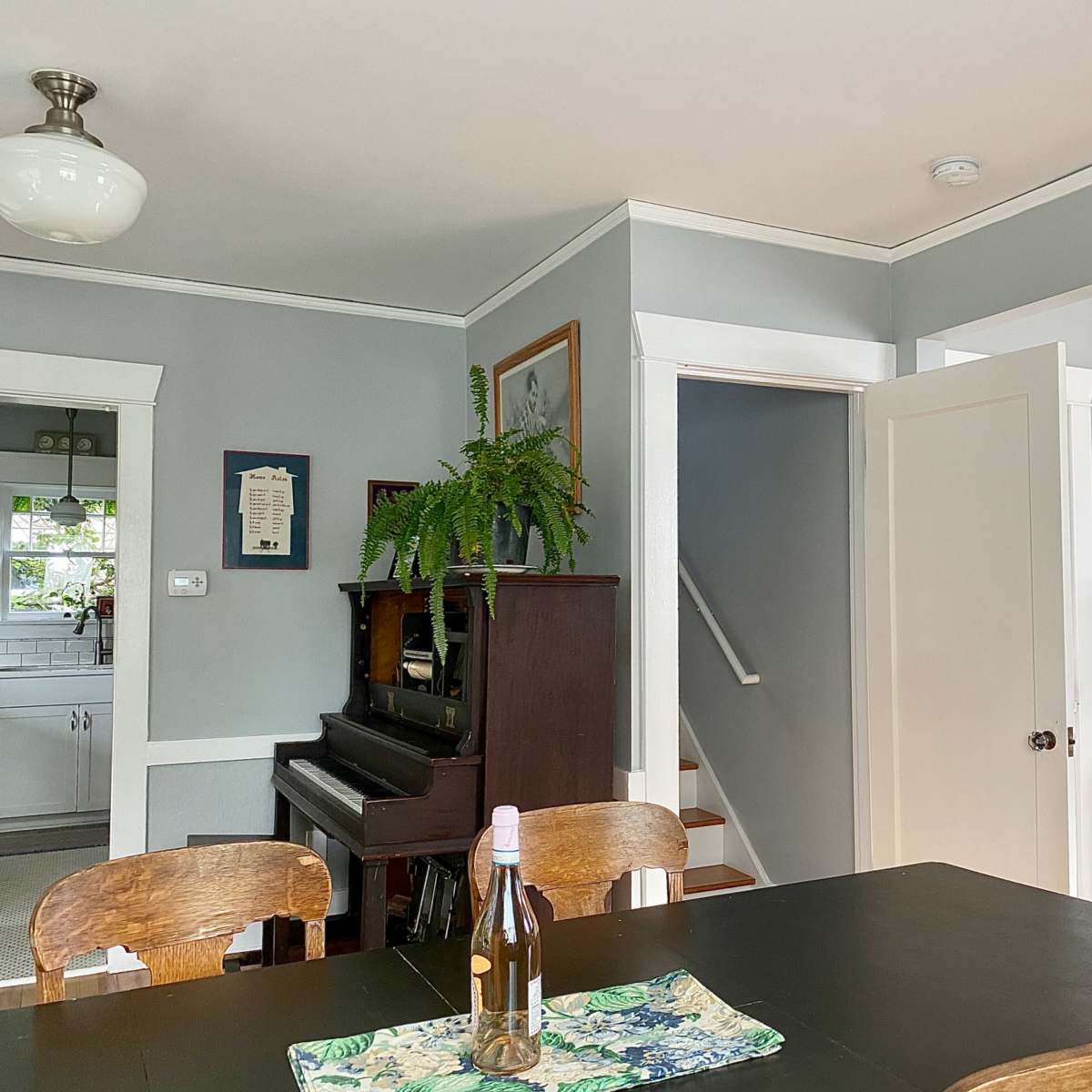 A dining area features a dark wooden table, a bottle of wine, and a piano near a staircase, with gray walls and a window leading to the kitchen.