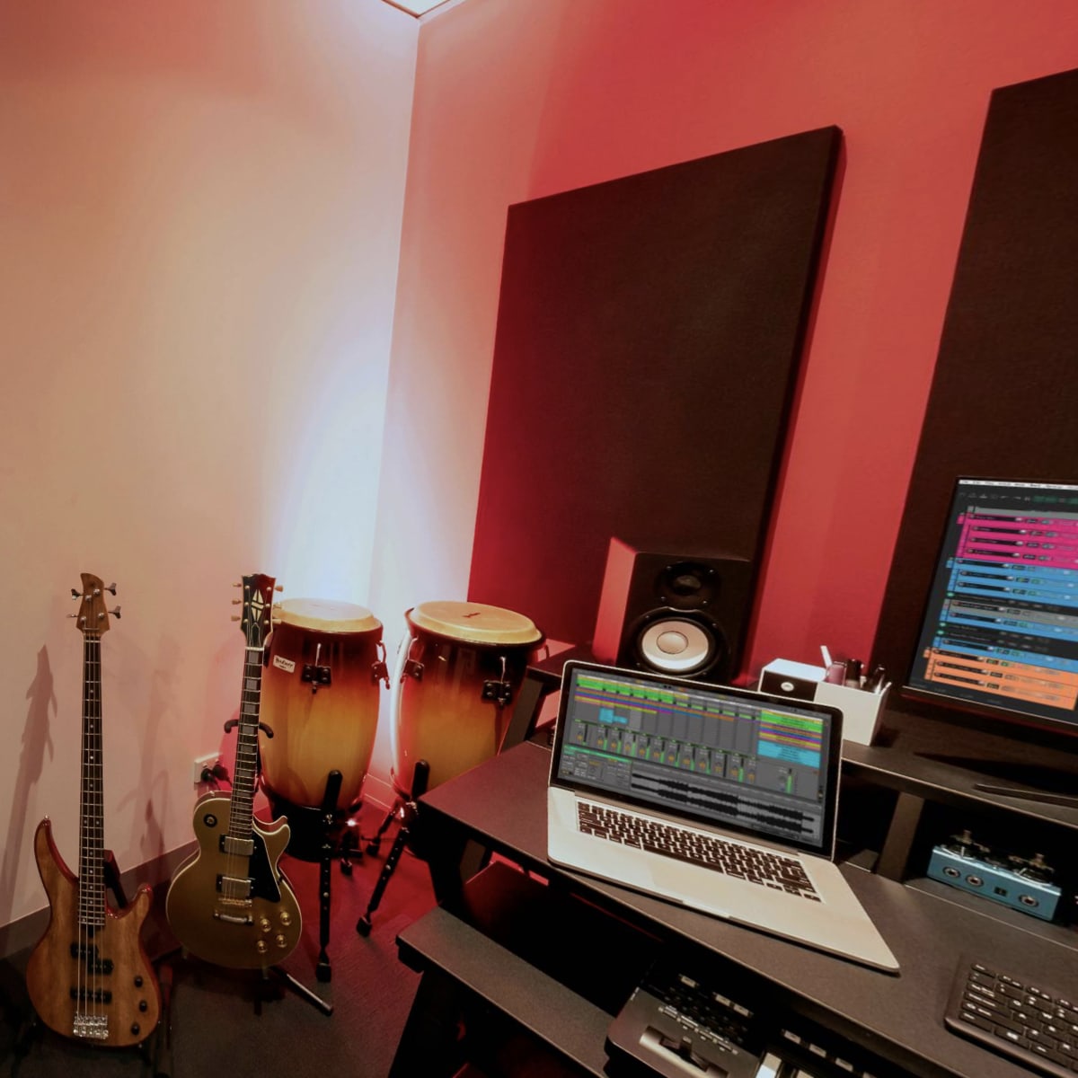 A music recording studio with a laptop on a desk, two congas, an electric bass guitar, and an amplifier against a red and white wall.