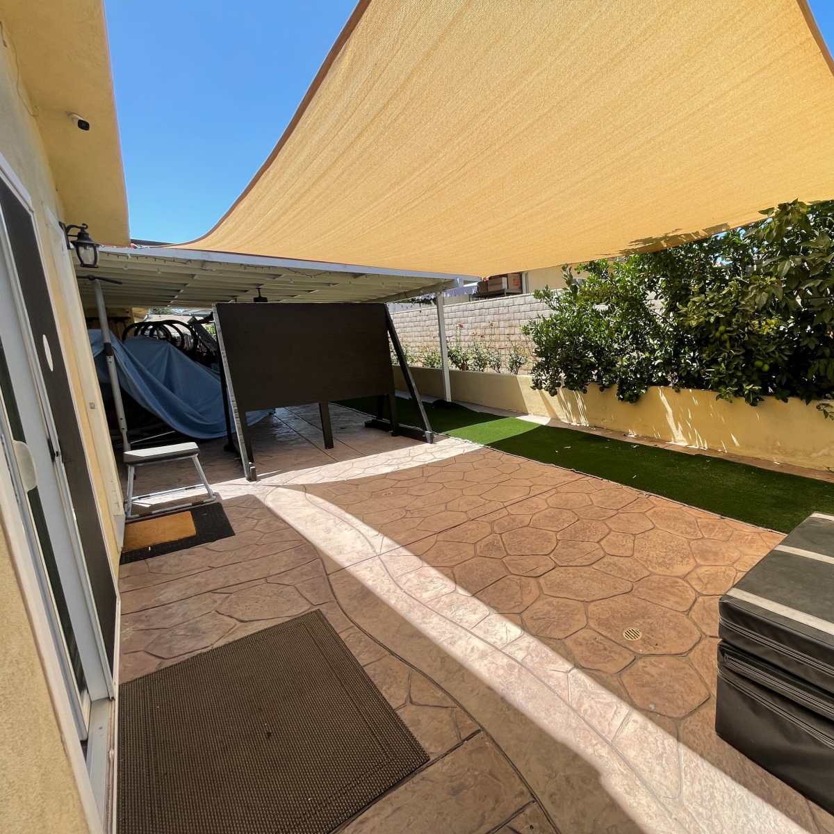 A patio area with a large shade sail overhead, featuring a textured concrete floor, artificial grass, and a covered storage space on one side.