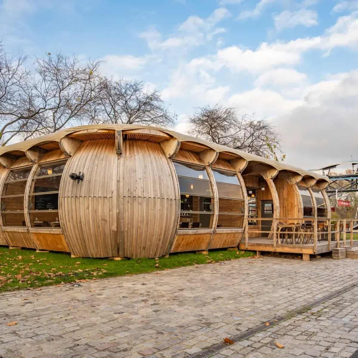 A wooden building with a curved design, featuring large windows and a porch area, situated on a stone pathway surrounded by trees.