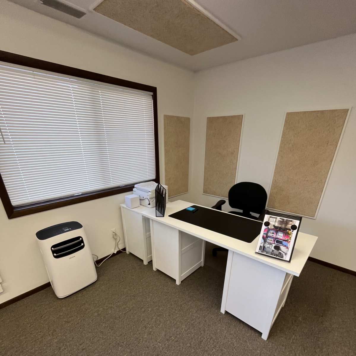 A minimalist office space featuring a white desk with a black chair, a file organizer, and an air conditioning unit against the wall.