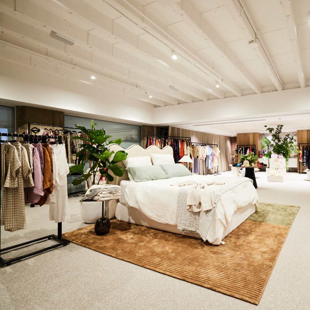 A spacious clothing store featuring a well-decorated display area with a bed, along with multiple racks of clothing and potted plants.