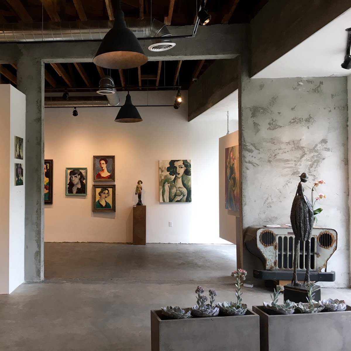 A modern art gallery featuring various paintings on the walls, a green bench, and sculptural displays in a spacious, well-lit environment with exposed ceilings and concrete flooring.