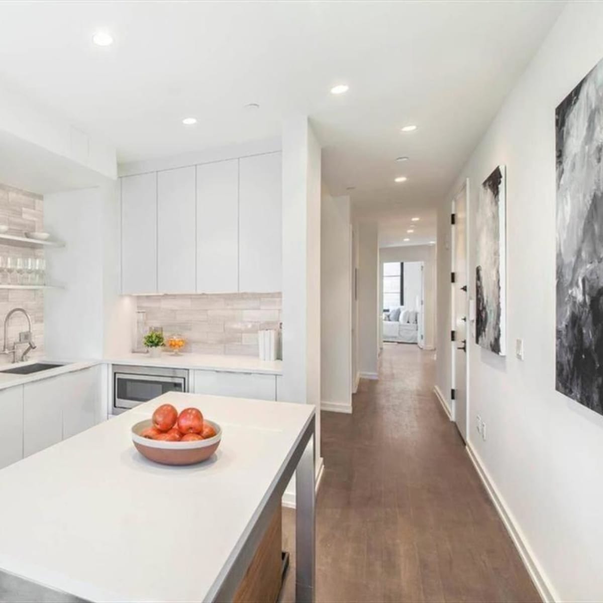 A modern kitchen features white cabinetry, a gray tiled backsplash, and a central island with a bowl of apples.