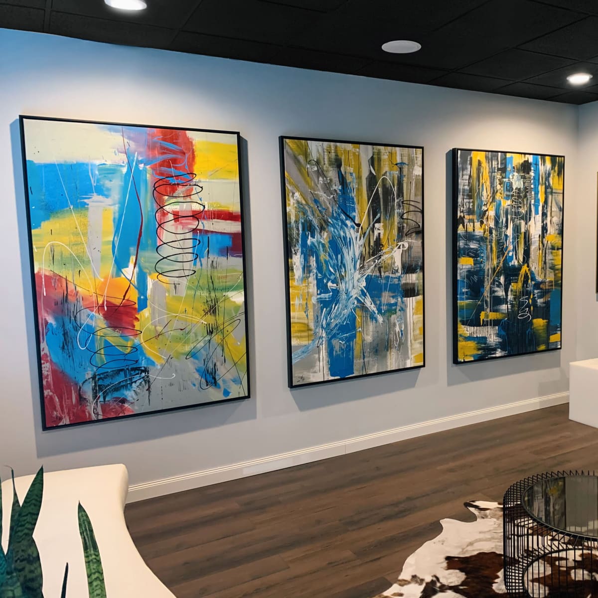 A modern art gallery with four colorful abstract paintings displayed on a light-colored wall, accompanied by a low round coffee table and a white pedestal.