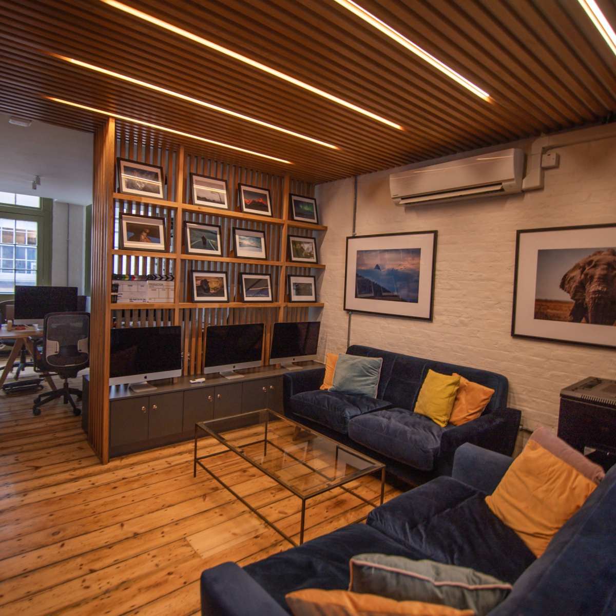 A modern office space with wooden flooring, featuring a cozy seating area with blue sofas and a wall of framed photos beside a workspace equipped with computers.