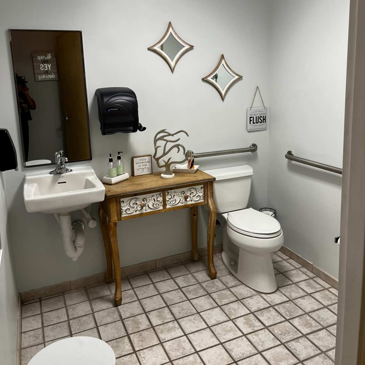 A restroom featuring a toilet, a sink with a mirror above it, decorative wall mirrors, and a small wooden table with various items on it.