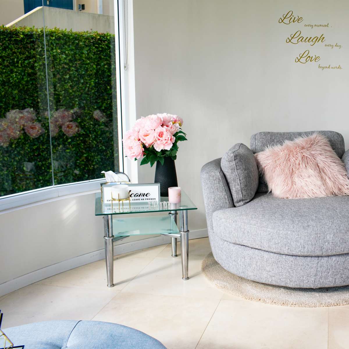A cozy living space with a gray circular chair, a glass side table, and pink flowers on the table, against a backdrop of green hedge and inspirational wall decor.