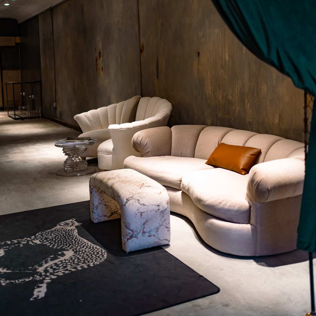 A modern seating area features a curved sofa and armchair, a marble bench, and a rug on a concrete floor, framed by green curtains.