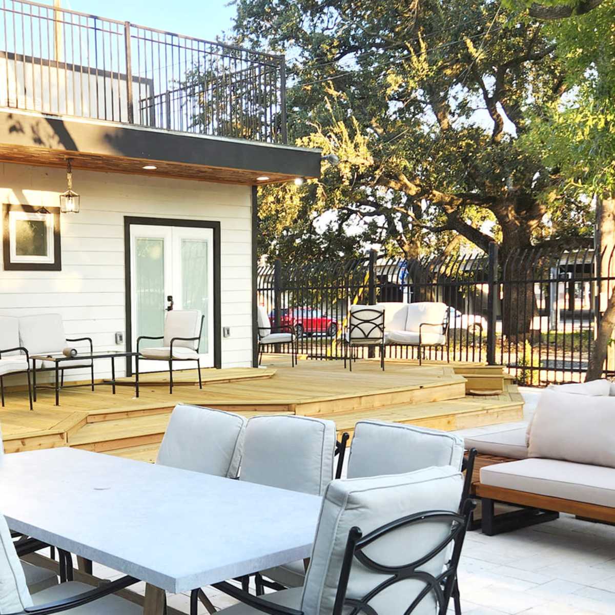 A patio area featuring a large dining table with chairs and a wooden deck surrounded by trees and a fenced area.
