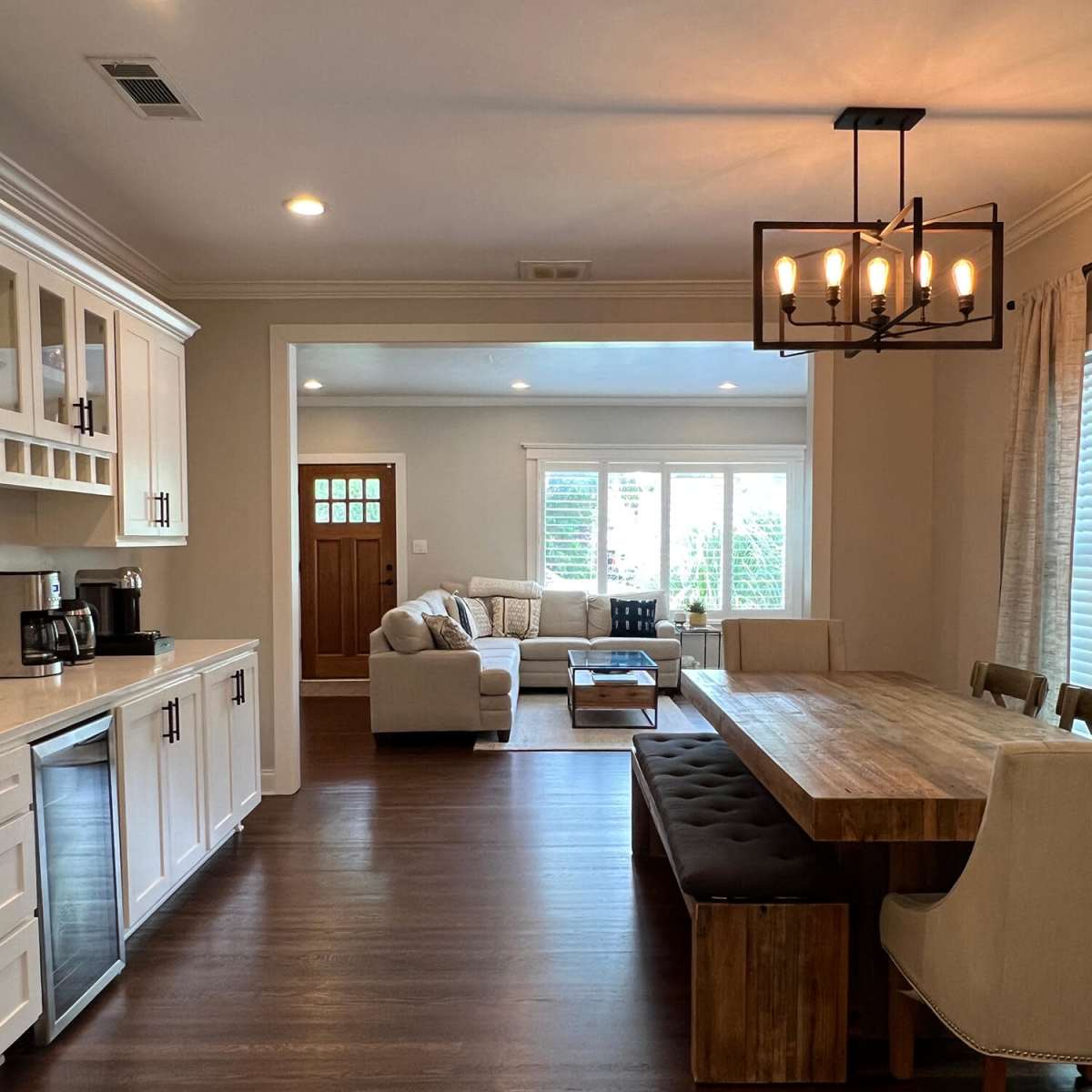 A modern kitchen and dining area features light wood cabinetry, a large wooden dining table, and a comfortable seating area with a sofa and windows providing natural light.